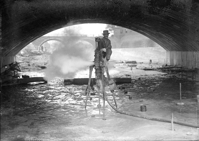 Here a workman poses with his steam drill under the Broad Street Bridge. Explosives will be used to deepen the river bed. [PHOTO: Albert R. Stone]