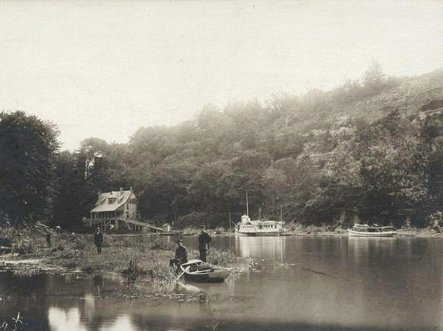 The Glen House, seen from river level, looking west. The scene shows a paddle wheel steam boat, the City of Rochester. A rowboat and men are along the bank of the river. [PHOTO: Rochester Public Library]