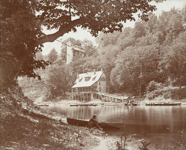 A view of the Glen House and its elevator along the banks of the Genesee River. In the foreground is a man in a straw hat seated by a rowboat.  [PHOTO: Rochester Public Library]
