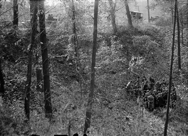 This distance shot shows the group of men gathered in the bottom of the gully. Along the top of the image can be seen the guard rails along the Webster State Road, men standing behind the rails, and vehicles parked along the road. [PHOTO: Albert R. Stone]