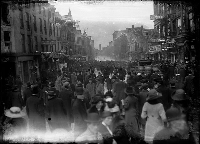 A large crowd is gathered on Front Street to look at the flood at the other end of the street. Varlan's Hotel, Rosenberg's Pawnshop, Charles Adam's Sons Grinding, J.G. Zweigle's Sons, W.T. Bridle Furniture, William Pigeon Shoes, Hyman Davis Shoes, Genesee Provision Company, Myers Department Store, and the F.H. Loeffler Company are visible in the photo. March, 1913. [IMAGE: Albert R. Stone]