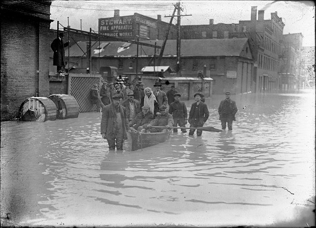 A group of people are pictured in and around a rowboat in the middle of flooded Front Street. They are members of a rescue crew that has saved a woman from a flooded building. All records for high water were broken in this flood. March, 1913. [IMAGE: Albert R. Stone]