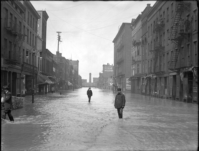 Men are standing ankle-deep in water on Front Street. On the left is the Atlantic Supply House and on the right is the People's Rescue Mission. March, 1913. [IMAGE: Albert R. Stone]