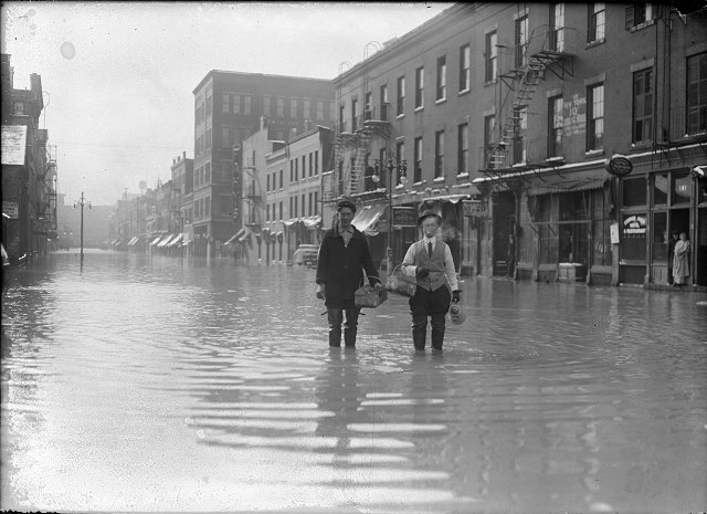 Two men stand ankle-deep in water on Front Street during the flood of 1913. They are carrying a coffee pot and baskets. On the right side of the photograph a man and a woman look out of a doorway. March, 1913. [IMAGE: Albert R. Stone]