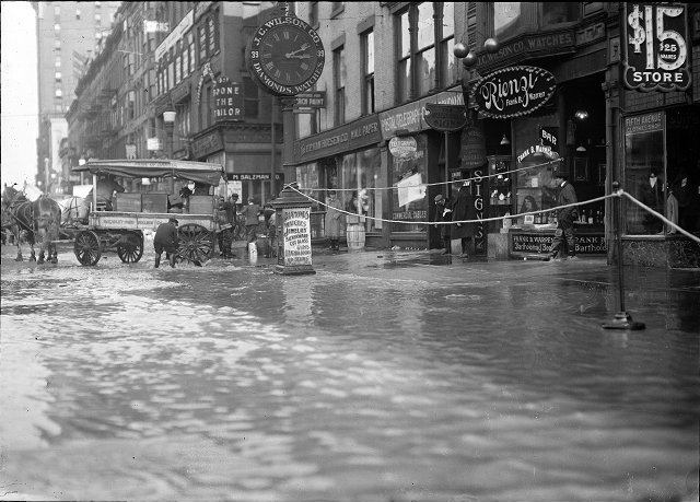 The flood has filled the store cellars with water on Main Street East at Graves Street. All the first floors are submerged as well. A horse and wagon are at the curb and the men are standing on the sidewalk. F.P. Van Hoesen Company, Rienzi (a bar), J.C. Wilson Company, the Postal Telegraph Cable Company, and the Fifth Avenue Clothes Shop are visible in the photo. March, 1913. [IMAGE: Albert R. Stone]