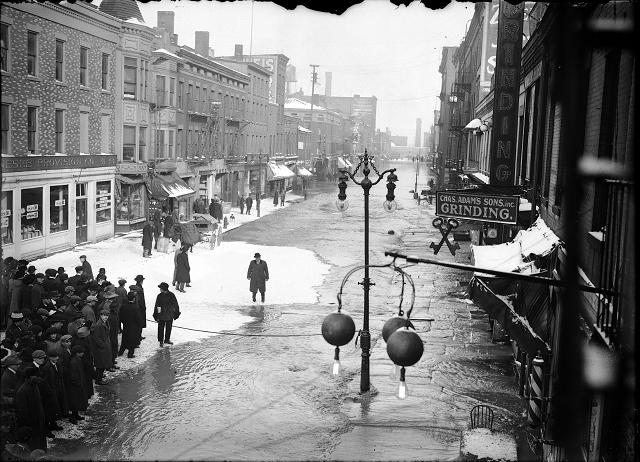 Rochesterians viewing the water on Front Street. Ice has formed in pockets on the flooded street. The sign for Charles Adam's Sons, Inc., 