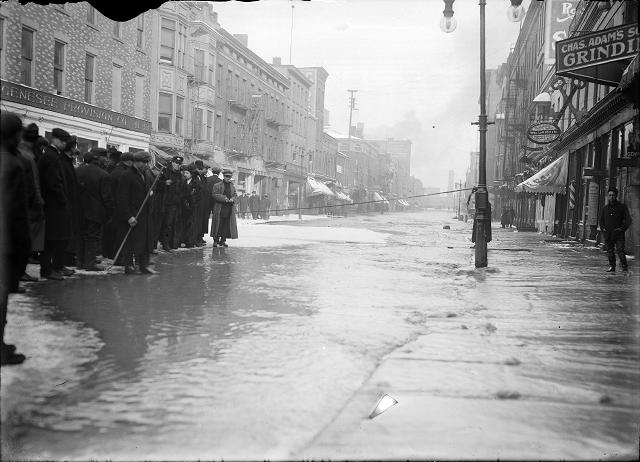 A group of people stand at the edge of the flood water on Front Street in front of the Genesee Provision Company. A rope cordons off the street near Charles Adam's Grinding and the William H. Larkin Hotel. March, 1913. [IMAGE: Albert R. Stone]
