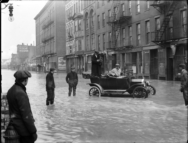Two men are stopped in a Ford car during the flood on Front Street. One man is holding a camera and other men stand in the street in ankle-deep water. March, 1913. [IMAGE: Albert R. Stone]