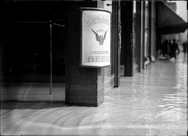 This view is a close-up of flood water going into the shops on Front Street. There is a Bartholomay Beer sign on the post. March, 1913. [IMAGE: Albert R. Stone]