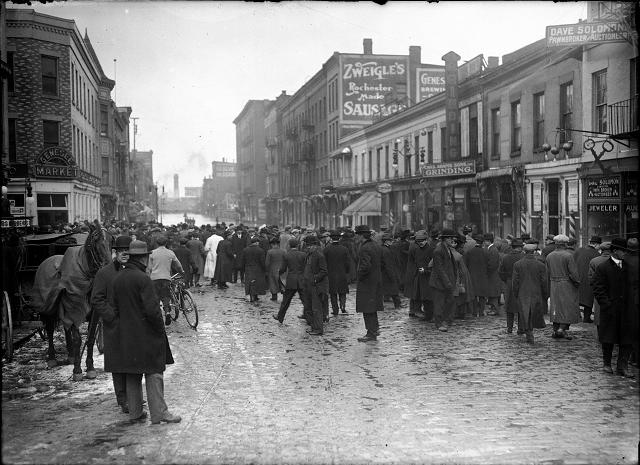 People, including one man with a horse and buggy, stand at the edge of the flood on Front Street. Dave Solomon, Pawnbroker and Auctioneer; Charles Adam's Sons, Incorporated, Grinding; Zweigle's; and (probably) the Genesee Provision Company are visible in the photo. March, 1913. [IMAGE: Albert R. Stone]