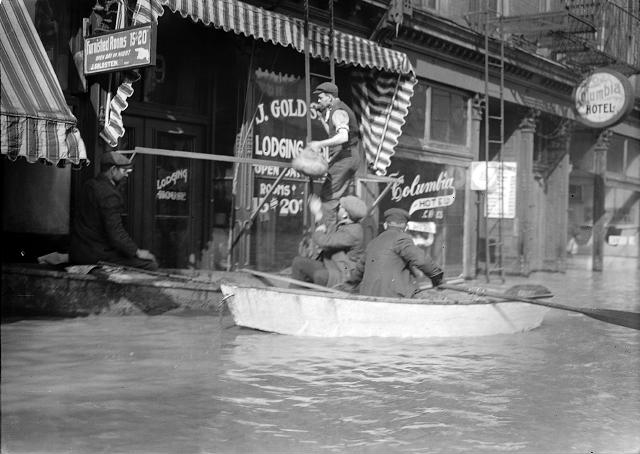 Men climb up a ladder from a rowboat to deliver food to families marooned on the upper floors of Rochester's business section. The men are pictured in front of Jacob Goldstein's Lodging House and J.C. Vine's Columbia Hotel. March, 1913. [IMAGE: Albert R. Stone]