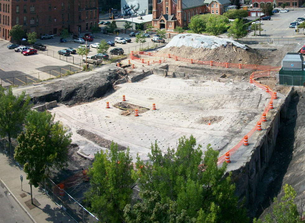 The auditorium floor and stage area of the old RKO Palace are on the left. The open pit on the right is filled with bricks and what appears to be a structural column. Mortimer Street is in the foreground with Saint Paul Street on the far left and Clinton Ave on the far right.