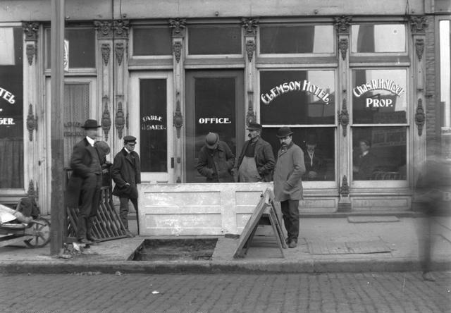These men are standing around a hole in the sidewalk where John Hornby and James Kenney fell through on December 11, 1901. In the background is the Clemson Hotel, with Charles R. Holliger, proprietor, at 52 South Avenue. [PHOTO: Rochester Municipal Archives]