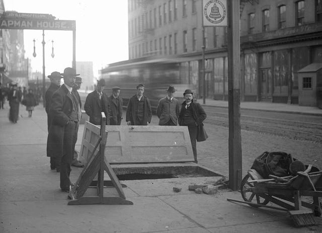 Looking south on South Avenue from just south of Ely Street. To the left is a sign for the Chapman House which was at 62 South Avenue, with Jay G. Longfellow as proprietor. Men are standing around a hole in the sidewalk where John Hornby and James Kenney fell through. A streetcar can be seen behind the men, and a wheelbarrow full of tools is in the foreground. A sign for Bell Telephone Company of Buffalo is on the post. [PHOTO: Rochester Municipal Archives]