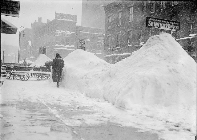 People walk along the snowy sidewalk on North Water Street after a snowstorm. This storm was 1914 (not the 1900 storm that Jim's Grandfather battled). The piles of snow between the sidewalk and the road are higher than people's heads. Sleds and wagons on sled runners are parked on the sidewalk. Visible business signs include Brewster, Gordon & Co., grocers, 39 North Water Street; Robertson & Sons, shoemakers, 38 North Water Street; Morrison Press, book and job printing, 60 North Water Street. [PHOTO: Rochester Municipal Archives]