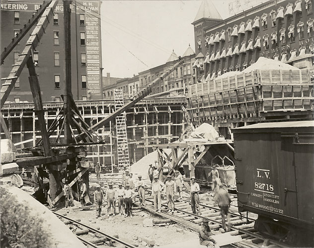Well-dressed men line up to climb a wooden ladder out of an excavation. The large area, bordered by steep stone walls, is part of the subway excavation. [Image from Albert R. Stone collection]