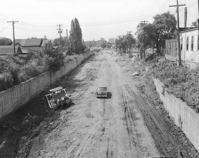 Construction of Interstate 490 in the Rochester subway bed along the former route of the Erie Canal. A bulldozer with a brush rake is doing the initial clearing of the site. The view is looking west from the Averill/Alexander Street area. July 1956.