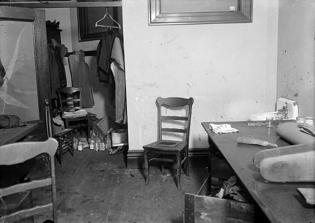 The floor of the closet in J. Frank O'Connor's tailor shop is full of gin bottles. Clothing under construction hangs in the closet. Several straight chairs stand around the room. The glass in the door to the room has been shattered. In the far right corner, beyond the table with a bolt of cloth, a pressing board, and a whisk broom, is the sink where the murderer supposedly washed his hands. [PHOTO: Albert R. Stone Collection]